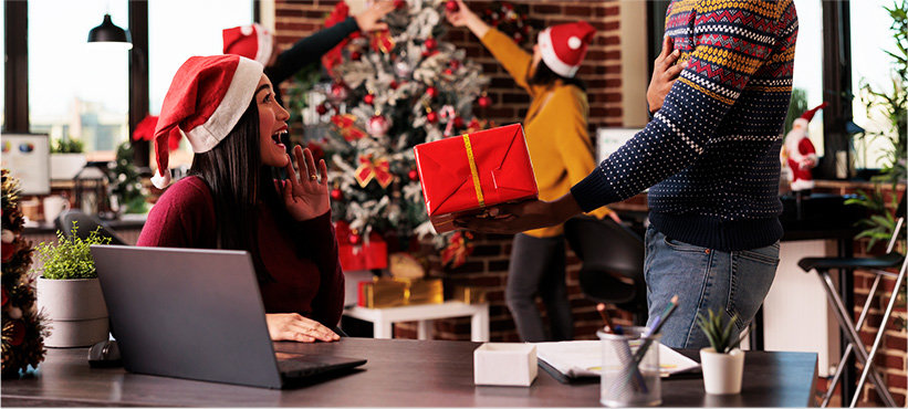Woman in holiday decorated corporate office looking surprised as co-worker hands her a gift.